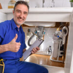 Plumbing technician checking water installation under the sink of a home kitchen with notepad and hand with ok gesture. Horizontal composition.